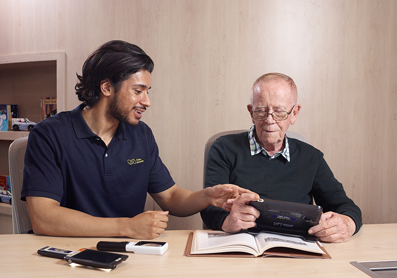 A Vision Australia staff member sitting at a table with an older client. A range of adaptive equipment is in front of them.